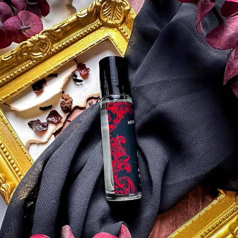 May include: A clear glass roller bottle with a black cap and a red and black label that says "DEVIL'S NIGHT". The bottle is sitting on a black fabric with a gold frame in the background.