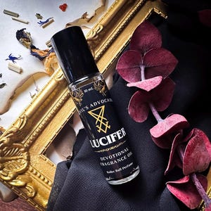 May include: A black rollerball perfume bottle with the text "Devil's Advocate Lucifer Devotional Fragrance Oil". The bottle has a black cap and a gold label with a sigil. The bottle is 10 ml.