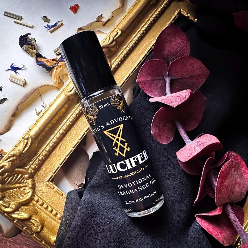 May include: A black rollerball perfume bottle with the text "Devil's Advocate Lucifer Devotional Fragrance Oil". The bottle has a black cap and a gold label with a sigil. The bottle is 10 ml.