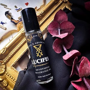 Puede incluir: Un frasco de perfume con bola rodante negro con el texto "DEVIL'S ADVOCATE" y "LUCIFER" en letras doradas. El frasco de 10 ml está sobre una tela negra, junto a un marco dorado y hojas burdeos.