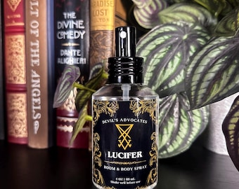 Pode incluir: Um frasco de spray de vidro transparente com uma tampa preta e um rótulo que diz "DEVIL'S ADVOCATES LUCIFER ROOM & BODY SPRAY". O rótulo tem detalhes dourados e um símbolo. A garrafa contém 118 ml de líquido.