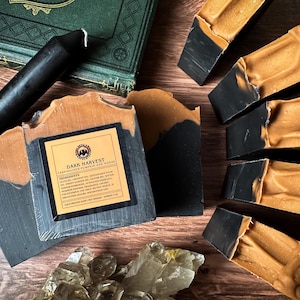 May include: A bar of handmade soap with a black and orange swirl design. The soap is labelled "Dark Harvest" with the scent "Caramelized Pumpkin and Woods". The soap is displayed on a wooden surface with a black candle and clear quartz crystals.