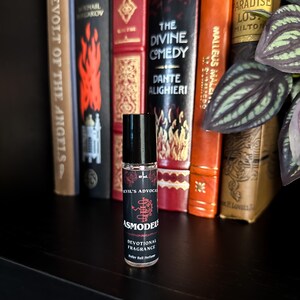 May include: A black rollerball perfume bottle with a red and white label. The label says "Evil's Advocate Asmodeus Devotional Fragrance Roller Ball Perfume". The bottle is sitting on a shelf with books behind it.