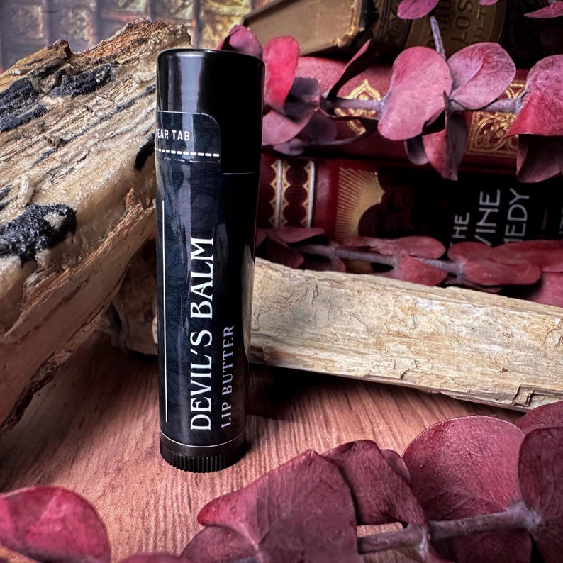 May include: A black lip balm tube with white text that reads "Devil's Balm Lip Butter". The tube is sitting on a wooden surface with a stack of books and red leaves in the background.