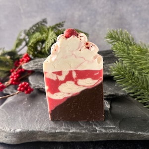 May include: A decorative soap bar with a layered design. The bottom section is dark brown, with a red and white marbled middle, topped with white swirls and a red berry. The soap is presented on a dark grey surface with sprigs of greenery.