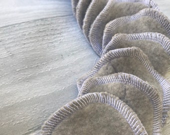 May include: A close-up of a pile of gray reusable makeup remover pads with white stitching around the edges.