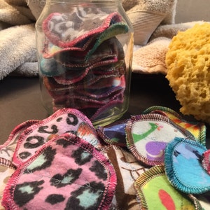 Puede incluir: Un tarro de vidrio lleno de discos de desmaquillante reutilizables. Los discos son de tela y tienen patrones coloridos. El tarro está sobre una superficie marrón.