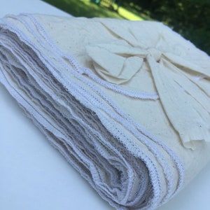 Op de afbeelding: Een stapel gevouwen, cr&egrave;mekleurige stoffen vierkanten met witte stiksels langs de randen. De stapel is vastgebonden met een bijpassende stoffen strik. De stof lijkt een natuurlijke vezel te zijn, mogelijk katoen of linnen.