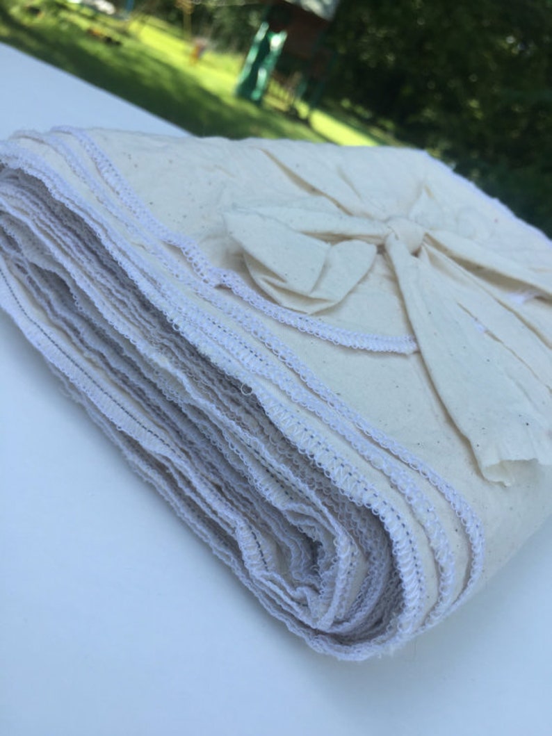 Op de afbeelding: Een stapel gevouwen, cr&egrave;mekleurige stoffen vierkanten met witte stiksels langs de randen. De stapel is vastgebonden met een bijpassende stoffen strik. De stof lijkt een natuurlijke vezel te zijn, mogelijk katoen of linnen.