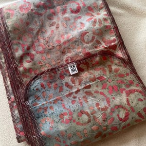 May include: Folded textile with a floral pattern in shades of pink, gray, and blue. The edges are finished with a dark burgundy stitch. A small white tag with black text is visible.