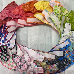 May include: A collection of colorful fabric baby cloth diapers with various patterns, including stars, flowers, and geometric designs. The diapers are arranged in a circle with the velcro closures facing outwards.
