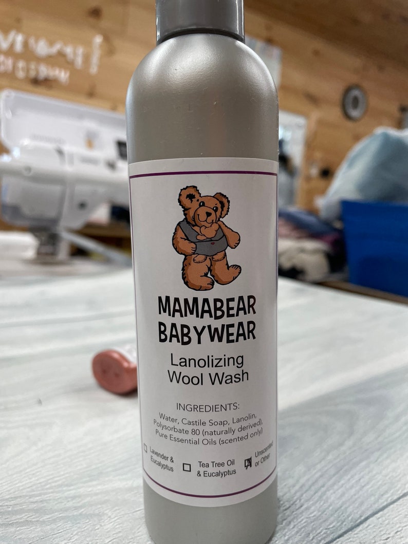 May include: A silver bottle of Mamabear Babywear Lanolizing Wool Wash with a cartoon bear wearing a vest on the label. The ingredients are listed on the label: Water, Castile Soap, Lanolin, Polysorbate 80 (naturally derived), Pure Essential Oils (scented only). The label also has checkboxes for Lavender & Eucalyptus, Tea Tree Oil & Eucalyptus, and Unscented or Other.