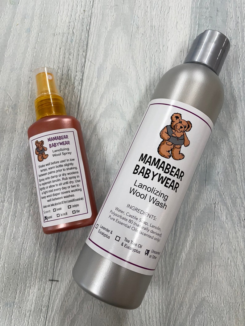 May include: Two bottles of Mamabear Babywear wool care products. A silver bottle of wool wash and a pink bottle of wool spray. Both bottles feature a cartoon bear logo and list ingredients and scent options.