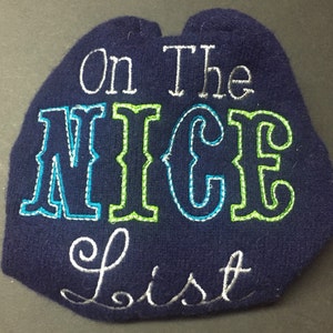 Pode incluir: Um chapéu de lã azul marinho com bordado branco que diz "On the Nice List". A palavra "Nice" é contornada por fio verde e azul.