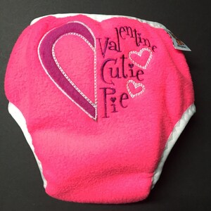 Puede incluir: Un pañal de bebé rosa brillante con un diseño de corazón y las palabras "Valentine Cutie Pie" bordadas en la parte delantera. El pañal tiene un ribete blanco y está hecho de un material suave y afelpado.