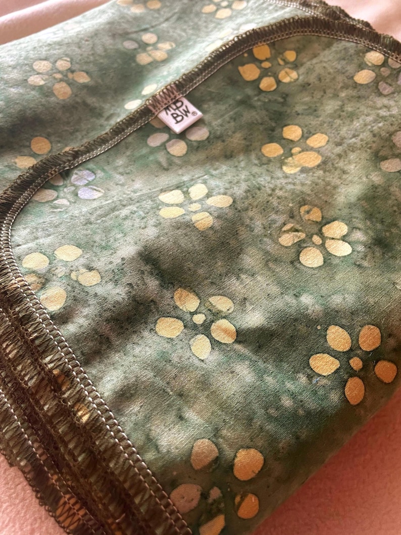 Op de afbeelding: Een groen en goud bloemenpatroon deken met een donkerbruine gestikte rand. De deken heeft een label met de tekst "MD BW". Het bloemenpatroon is een herhalend ontwerp van gele bloemen op een groene achtergrond.