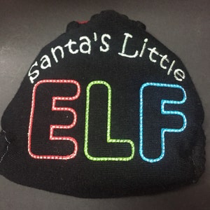 Pode incluir: Chapéu de malha preto com bordado branco que diz "Santa's Little Elf". As letras são contornadas em vermelho, verde e azul.