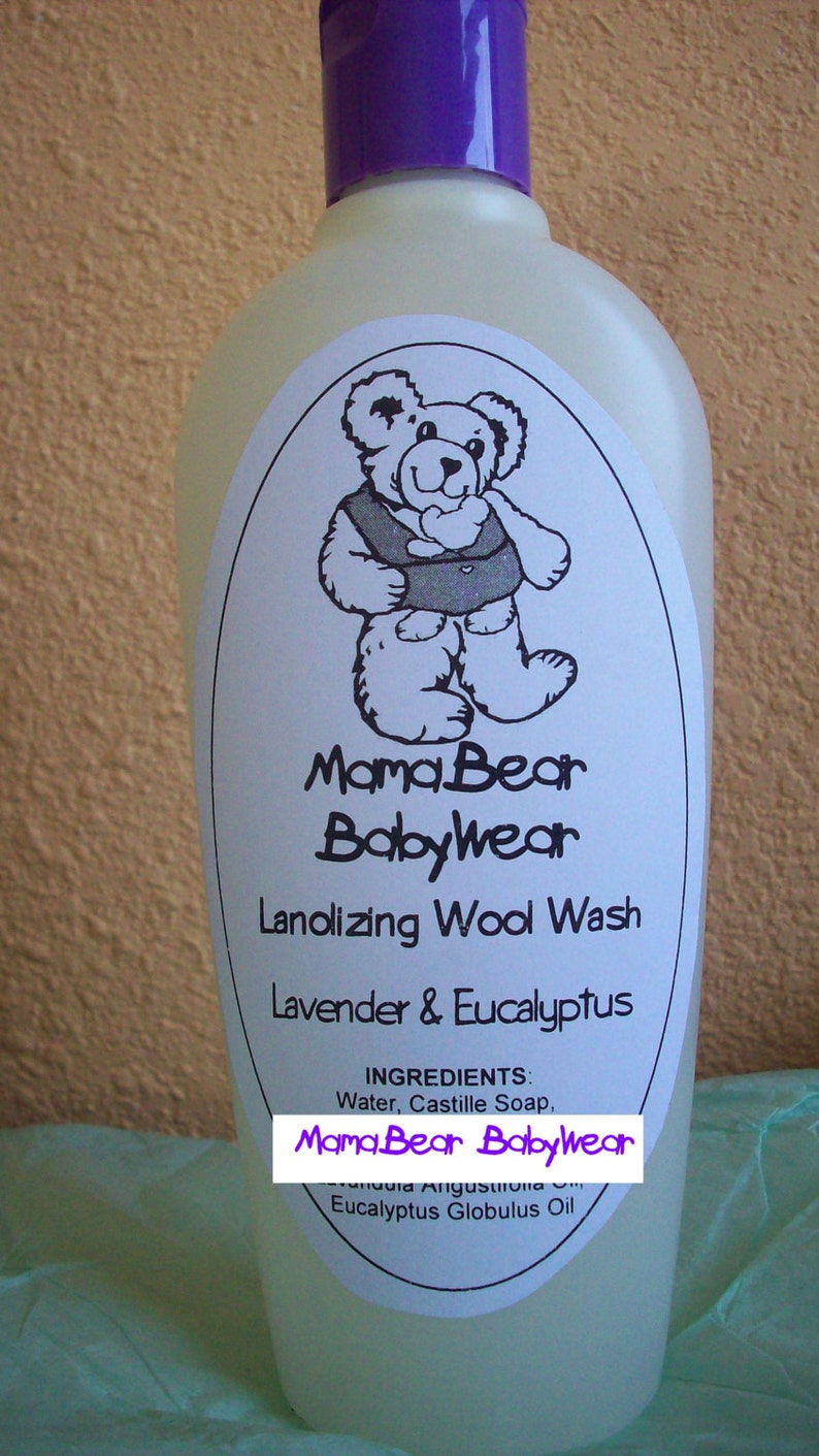 May include: A clear plastic bottle with a purple lid and a white label. The label features a cartoon bear wearing a baby carrier and the text "Mama Bear Babywear Lanolizing Wool Wash Lavender & Eucalyptus Ingredients: Water, Castille Soap, MamaBear Babywear Eucalyptus Globulus Oil"