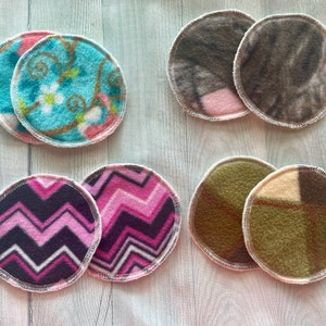 May include: Six round fabric nursing pads in various patterns. Two pads feature a blue and pink floral design, two pads feature a pink and black chevron pattern, and two pads feature a green and brown plaid pattern.