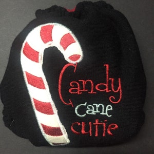 Pode incluir: Capa de fralda de tecido preto com uma bengala de doces vermelha e branca e o texto "Candy Cane Cutie" bordado.