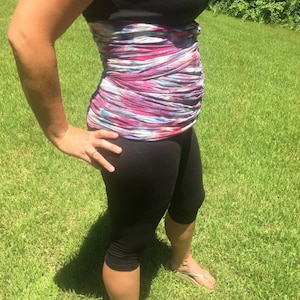 Op de afbeelding: Een persoon draagt een zwarte tanktop met een kleurrijk, patroon middenrif en zwarte capri leggings. De persoon staat op groen gras in het zonlicht en draagt teenslippers.