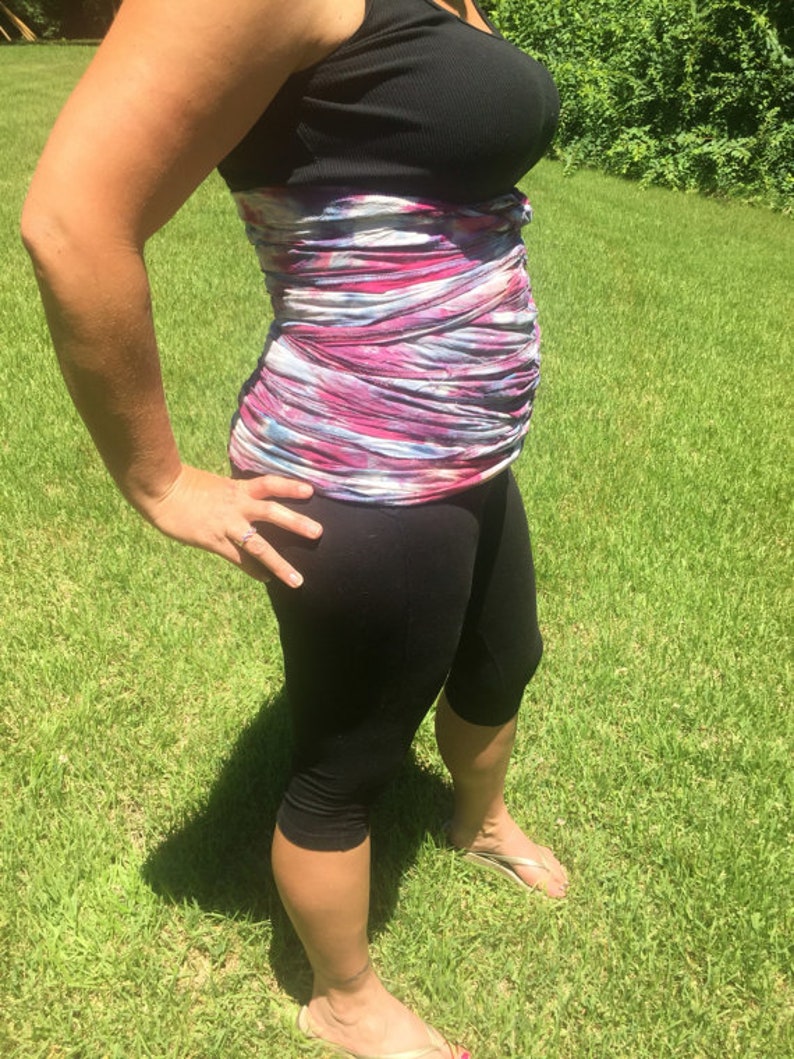 Op de afbeelding: Een persoon draagt een zwarte tanktop met een kleurrijk, patroon middenrif en zwarte capri leggings. De persoon staat op groen gras in het zonlicht en draagt teenslippers.
