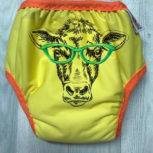 Puede incluir: Pañal de tela reutilizable amarillo con una imagen bordada de una vaca con gafas verdes.