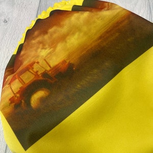 May include: A yellow cloth diaper with a printed image of a tractor in a field. The tractor is brown and yellow and is driving through a field of green grass. The sky is a bright orange and yellow.