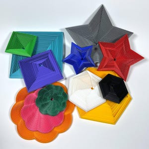 May include: A collection of colorful, geometric 3D-printed shapes. The shapes include stars, squares, hexagons, and a flower. Colors include green, blue, purple, red, gray, white, black, yellow, orange, and pink. The shapes are arranged on a white background.