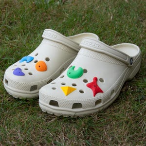 Könnte beinhalten: Ein Paar beigefarbene Crocs-Clogs mit bunten, geometrischen Jibbitz-Anhängern. Die Anhänger umfassen blaue, orange, grüne, gelbe und rote Formen. Die Clogs stehen auf grünem Gras.