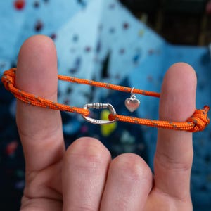 Peut inclure: Bracelet en corde orange et grise avec un mousqueton argenté et un petit charme en forme de cœur argenté. Le bracelet est attaché autour d'un doigt.