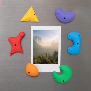 Peut inclure: Ensemble de prises d'escalade colorées et une photographie de paysage encadrée, disposées sur une surface grise. Les prises d'escalade sont de formes et de couleurs variées : jaune, rouge, violet, bleu, orange et vert. La photographie représente une chaîne de montagnes.