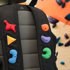 Peut inclure: Un sac à dos noir avec un panneau dorsal rembourré gris portant l'inscription Tahoe BP 150. Les bretelles sont ornées de six prises d'escalade colorées en rouge, jaune, bleu, orange, violet et vert, chacune avec des trous de vis.