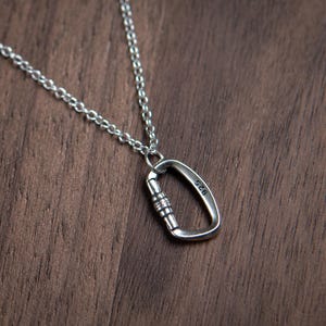 Peut inclure: Un pendentif mousqueton en métal argenté est suspendu à une chaîne assortie de couleur argentée. Le mousqueton présente un mécanisme de fermeture fonctionnel et porte l'inscription 925 sur le côté, indiquant une composition en argent sterling.