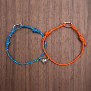 May include: Two matching bracelets with blue and orange rope, each with a silver clasp. One bracelet has a silver heart charm. The bracelets are displayed on a wooden surface.