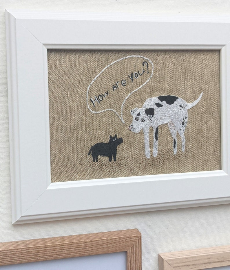 May include: Embroidered artwork on burlap fabric in a white frame. The artwork features two dogs, one black and one white with black spots, with a speech bubble above the white dog that says "How are you?"