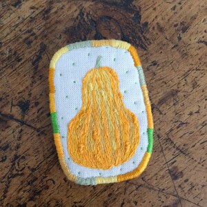 Puede incluir: Un parche bordado a mano que presenta una calabaza amarilla con un tallo verde. El parche está rodeado por un borde multicolor con acentos verdes, amarillos y naranjas.