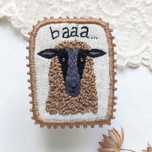 Puede incluir: Broche bordado de oveja con la palabra "baaa" sobre la cabeza de la oveja. La oveja tiene un abrigo lanudo marrón, cara negra y orejas negras. El broche está enmarcado con un borde de cuentas marrón.