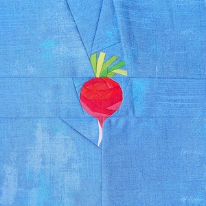 May include: A fabric quilt block featuring a central red radish appliqué with green leaves, constructed from multiple small fabric pieces. The radish is set against a solid blue background with a subtle mottled texture and visible seams.
