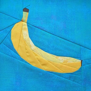 Könnte beinhalten: Ein Stoff-Quiltblock mit einem gelben Bananenmotiv, das in Paper-Piecing-Technik gefertigt wurde. Die Banane ist vor einem leuchtend blauen Hintergrund platziert, der aus mehreren winkligen Stoffsegmenten mit sichtbaren Nähten besteht.