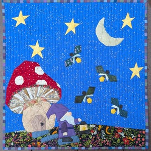 Puede incluir: Un edredón hecho a mano que representa una escena nocturna con una seta roja con lunares blancos, un gnomo, estrellas, una luna creciente y criaturas voladoras sobre un fondo azul. El edredón tiene un borde floral y está acolchado a máquina.