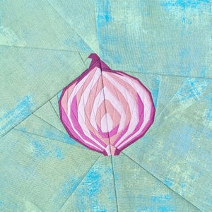 May include: A fabric quilt block featuring a central pieced onion design in shades of purple, magenta, and light pink. The onion is set against a background of light green and blue mottled fabric, constructed with straight seams and geometric shapes.