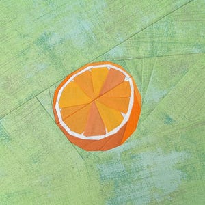May include: A fabric quilt block featuring a pieced orange slice motif in shades of orange and yellow with a white rind. The citrus slice is centered on a textured light green background with visible seam lines from the paper piecing technique.