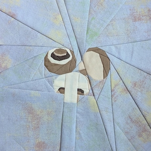 May include: A fabric quilt block featuring a foundation paper pieced design. The pattern displays a central light beige and brown geometric shape surrounded by radiating triangular segments of light blue fabric with a mottled, textured appearance.
