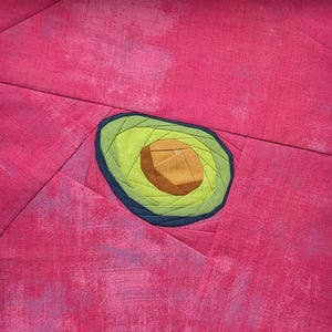 May include: A fabric quilt block featuring a central avocado motif constructed from layered green and brown fabric pieces. The avocado is surrounded by a dark blue border and set against a background of textured, mottled hot pink fabric.