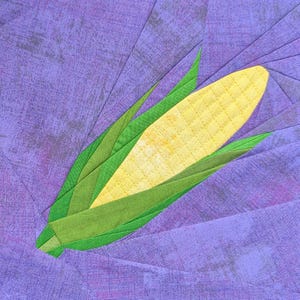 May include: A fabric quilt block featuring a central yellow corn cob with a quilted grid pattern, surrounded by layered green fabric husks. The entire design is set against a background of purple fabric pieces arranged in a radiating pattern.