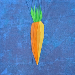May include: A fabric quilt block featuring a central carrot motif constructed from orange and yellow geometric fabric pieces with bright green fabric leaves, all set against a textured, mottled dark blue fabric background.
