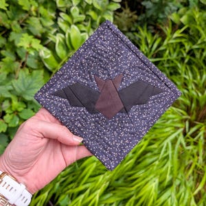 Puede incluir: Un posavasos cuadrado y acolchado con un diseño de murciélago. El posavasos tiene un fondo azul oscuro con patrones de estrellas blancas. El murciélago está hecho de tela negra y marrón, añadiendo un toque de decoración de Halloween.