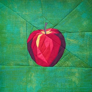 Puede incluir: Un bloque de edredón de tela con un diseño de manzana hecho con la técnica de paper piecing en tonos rojo, morado oscuro y amarillo. La manzana está centrada sobre un fondo de tela verde texturizada con costuras geométricas que irradian.