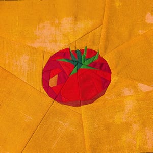May include: A fabric quilt block featuring a central red tomato with a green stem, constructed from multiple geometric fabric pieces. The tomato is set against a background of golden yellow fabric panels arranged in a radiating sunburst pattern.